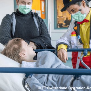 Ein Kind auf einer Krankenhausliege lächelt einem Clown mit Maske zu, während eine Begleitperson im Hintergrund steht.