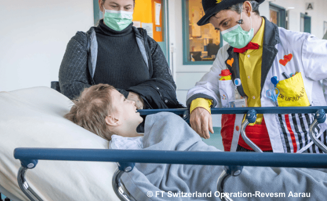 Ein Kind auf einer Krankenhausliege lächelt einem Clown mit Maske zu, während eine Begleitperson im Hintergrund steht.