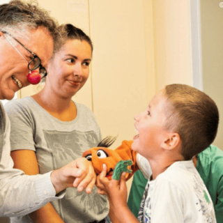 Ein Clown mit roter Nase bringt ein lachendes Kind mit einer Handpuppe zum Lachen, während zwei Erwachsene dabei zusehen.