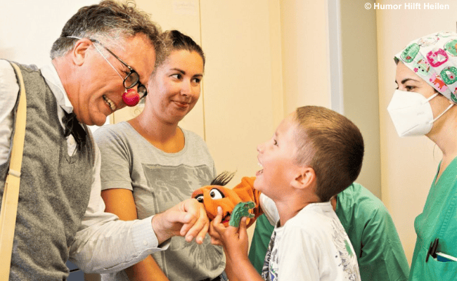 Ein Clown mit roter Nase bringt ein lachendes Kind mit einer Handpuppe zum Lachen, während zwei Erwachsene dabei zusehen.