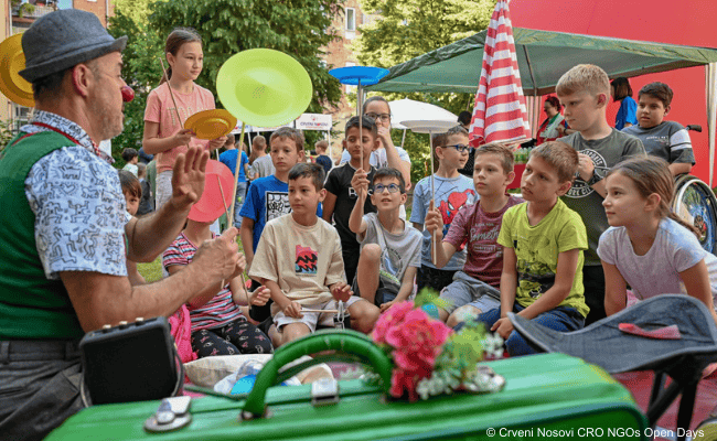 Ein Clown mit roter Nase führt eine Jonglier-Show mit Tellern vor, während Kinder begeistert zusehen und selbst üben.