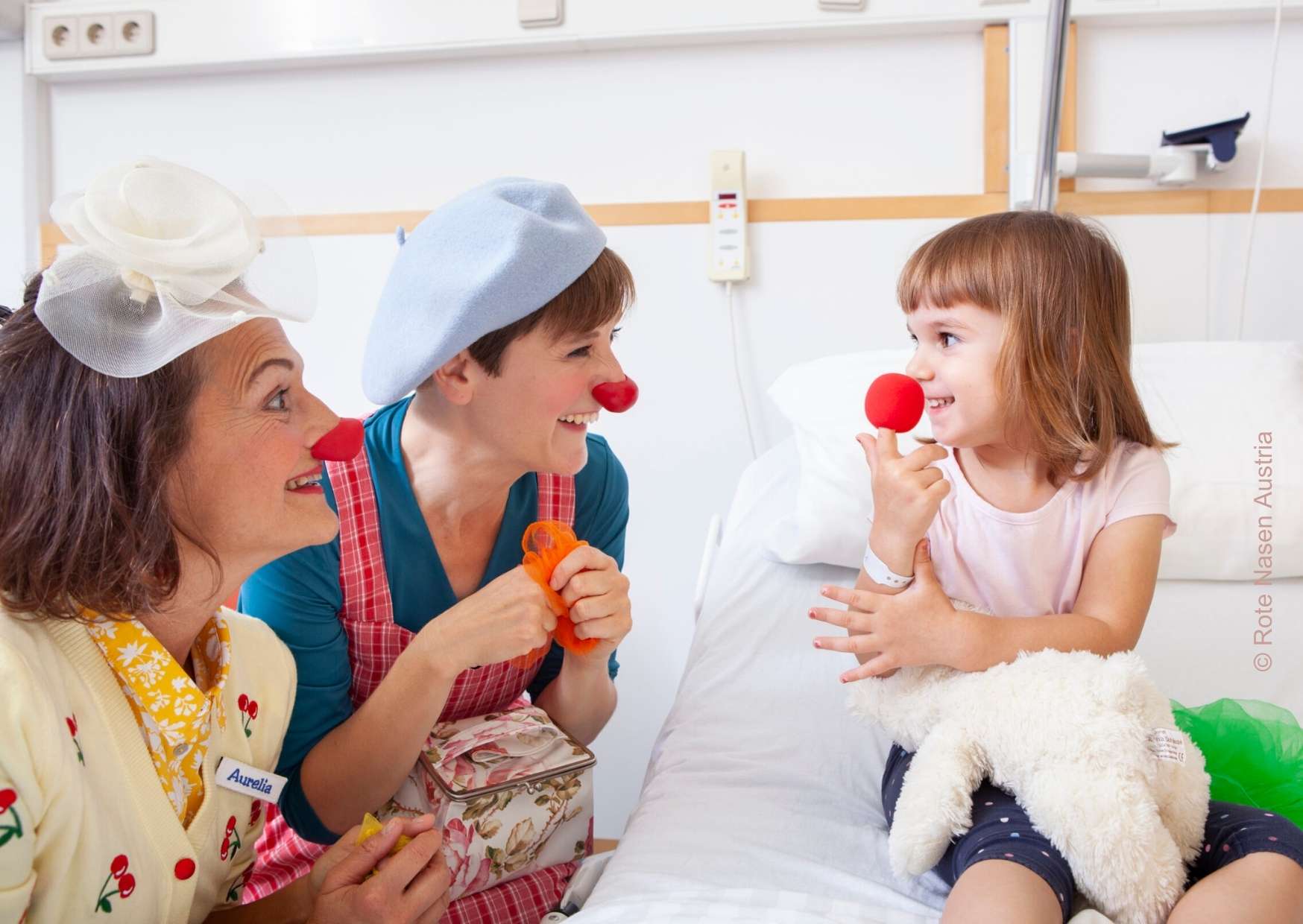 Zwei Clowns bringen ein Mädchen im Krankenhausbett mit roten Nasen und spielerischem Charme zum Lachen. Freude im Krankenzimmer.