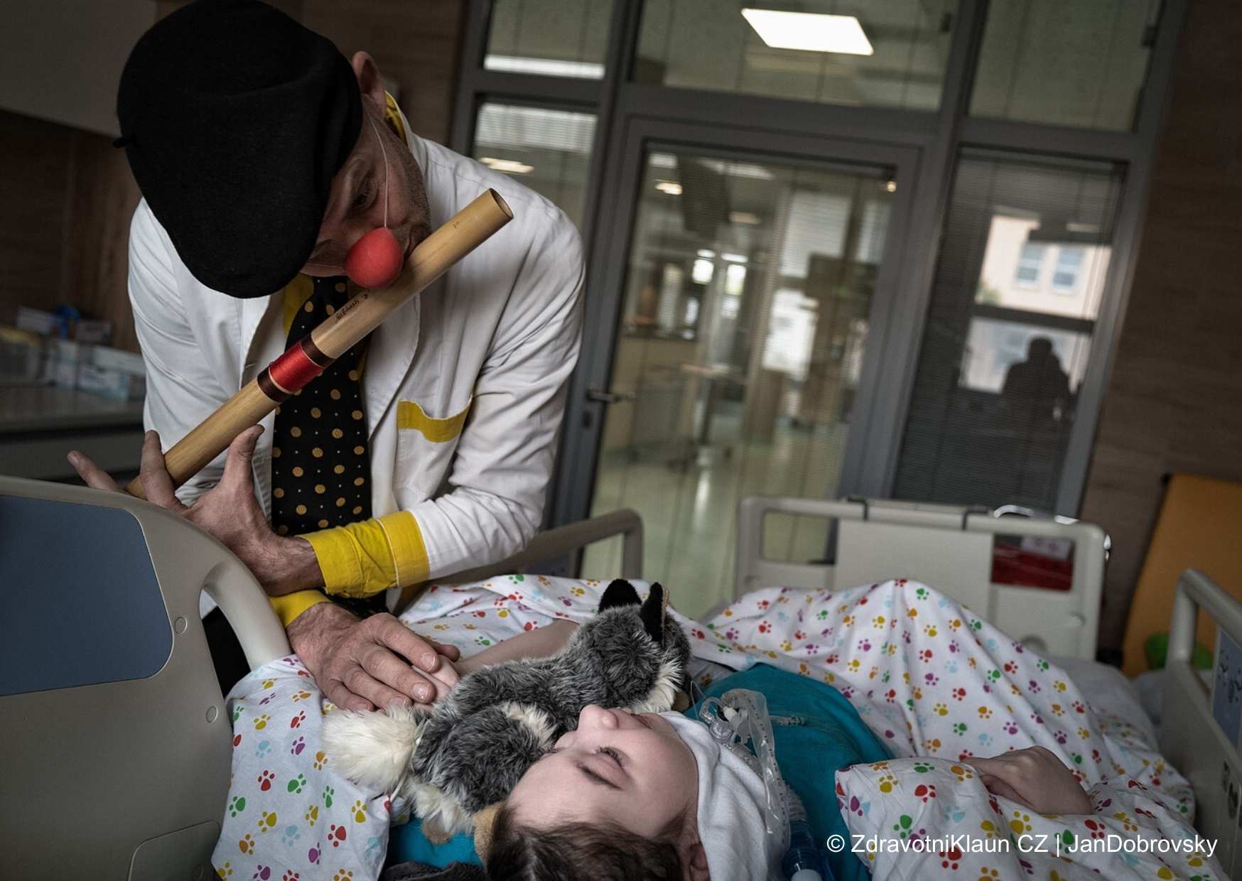 Ein Clown im Krankenhaus spielt auf einer Flöte für ein Kind, das in seinem Bett liegt und ein Stofftier liebevoll umarmt.