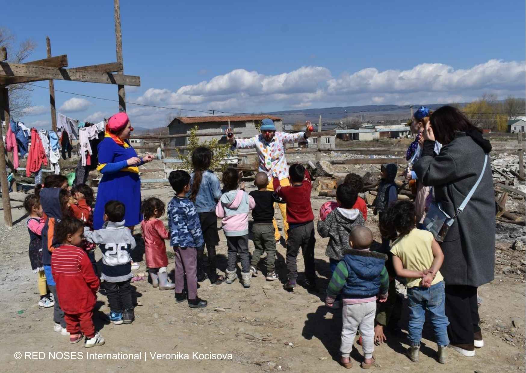 Ein Clown führt eine fröhliche Show für eine Gruppe von Kindern in einer ländlichen Gegend auf, umgeben von einfachen Behausungen.