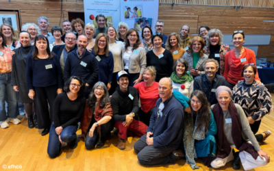 Eine große Gruppe von Menschen posiert lächelnd für ein Gruppenfoto in einem Holzsaal mit Bannern im Hintergrund..