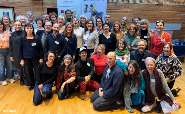 Eine große Gruppe von Menschen posiert lächelnd für ein Gruppenfoto in einem Holzsaal mit Bannern im Hintergrund..