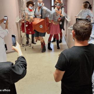 Clowns führen eine humorvolle Parade in einem Krankenhausflur auf, während Personal und Besucher die Szene mit Handys filmen.