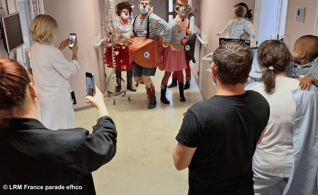 Clowns führen eine humorvolle Parade in einem Krankenhausflur auf, während Personal und Besucher die Szene mit Handys filmen.