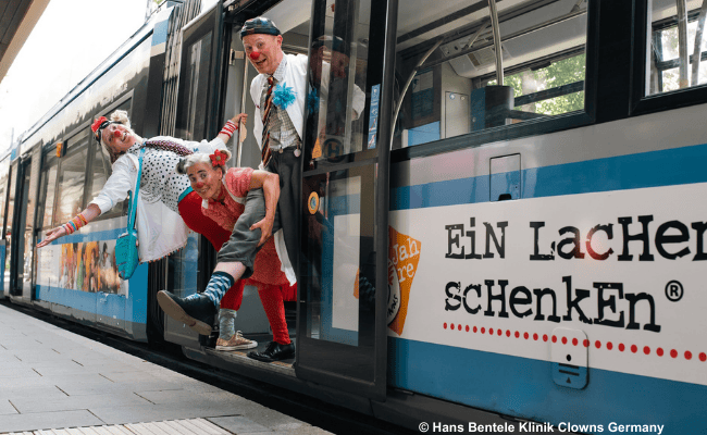 Drei Klinikclowns posieren spielerisch an einer Straßenbahn mit der Aufschrift 'Ein Lachen schenken' und sorgen für Heiterkeit.