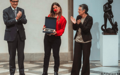 Eine Frau mit roter Clownsnase hält eine Auszeichnung, während zwei Personen daneben applaudieren.