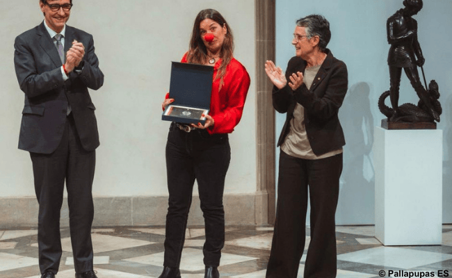 Eine Frau mit roter Clownsnase hält eine Auszeichnung, während zwei Personen daneben applaudieren.